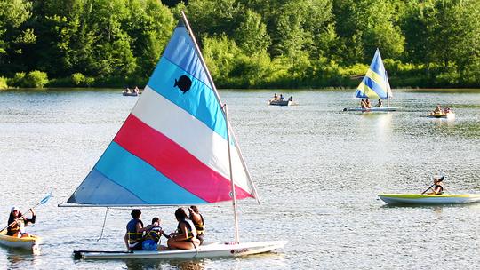 A variety of paddle boats and sail boats with candy striped sails cover the dark green surface of Lake Arax at Camp Nubar.